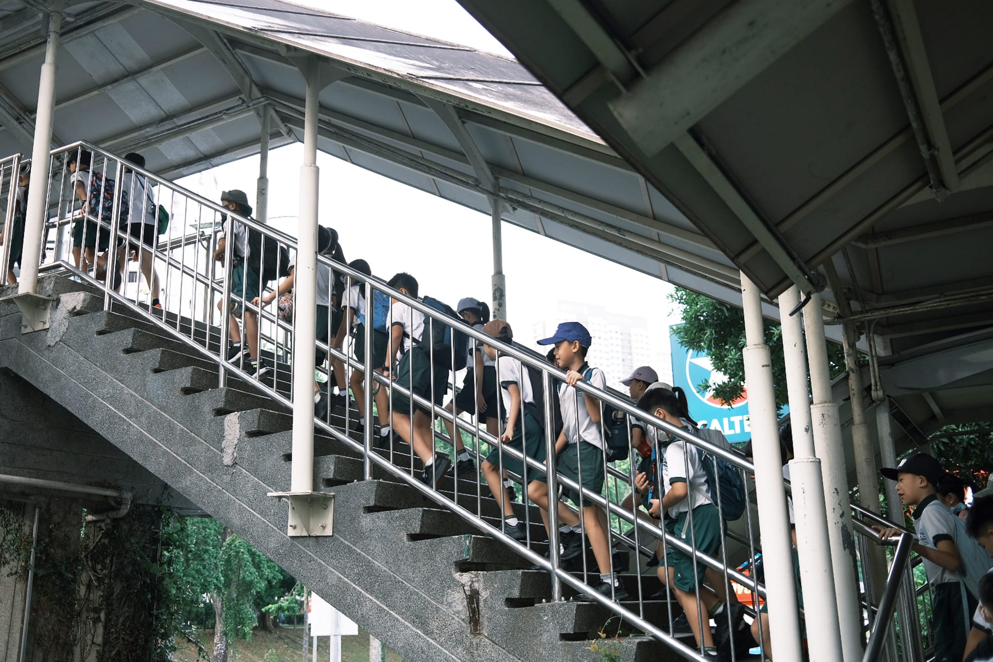 Primary 4 students wearing school uniforms, caps, and backpacks walk together up a pedestrian overhead bridge during their Resilience Walk.